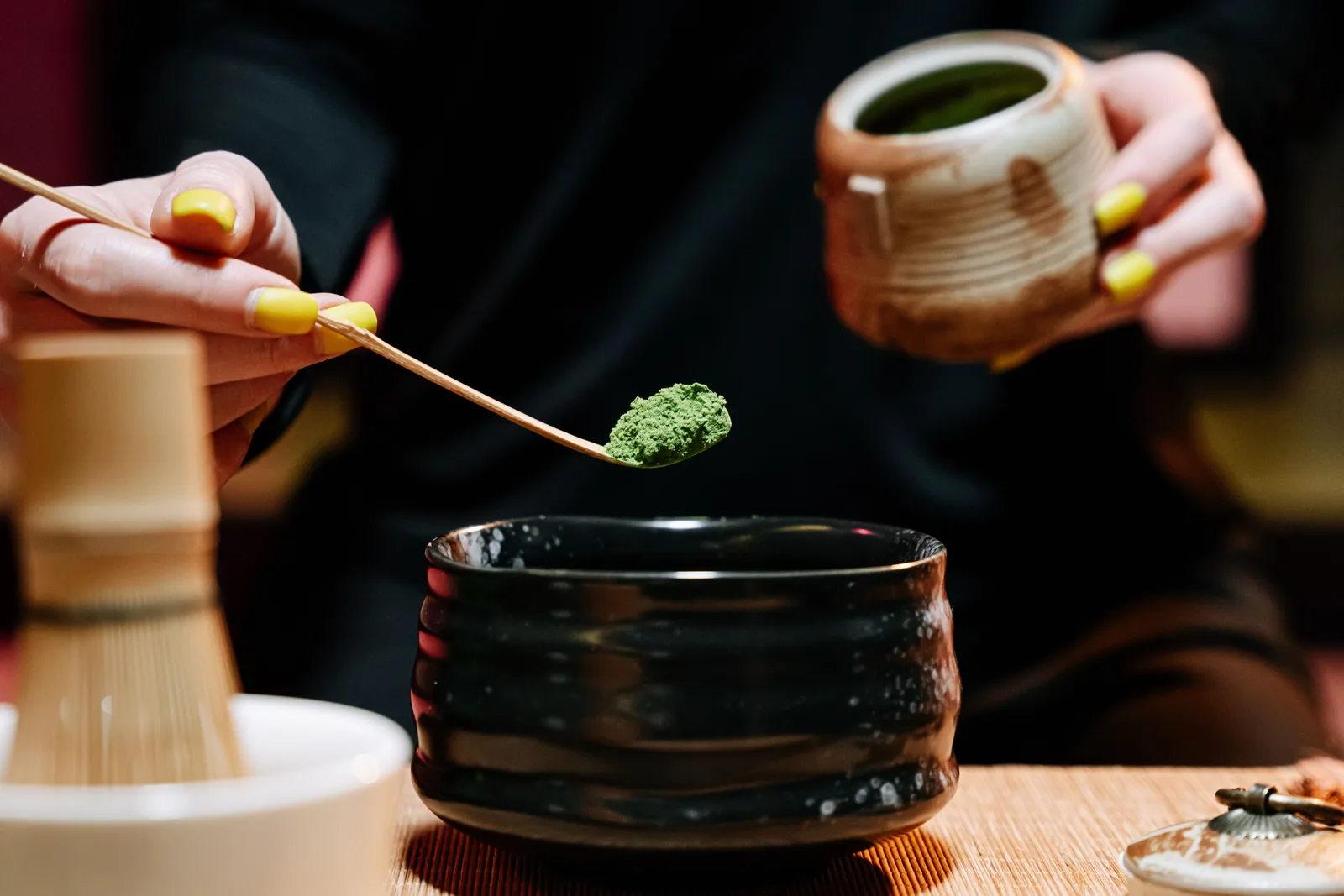 Matcha powder being measured with a bamboo scoop into a ceramic bowl beside a chasen whisk